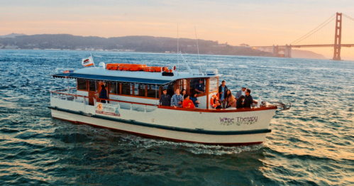 A boat named "Wine Therapy" with passengers cruising on water near the Golden Gate Bridge at sunset.