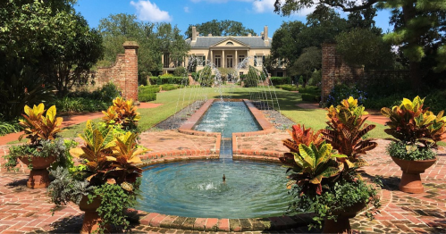 A lush garden with a fountain, colorful plants, and a grand house in the background under a clear blue sky.