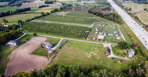 Aerial view of a rural area with fields, a maze, and tents near a highway, surrounded by trees and farmland.