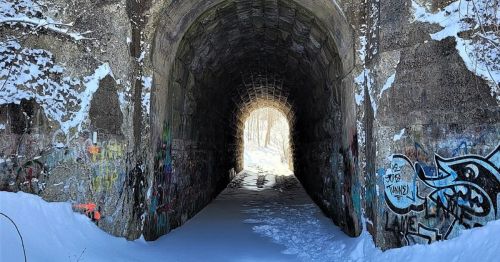 A snow-covered tunnel with graffiti on the walls, leading to a bright light at the end.