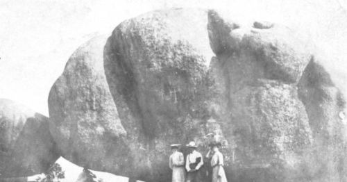 Three people stand in front of a large boulder, surrounded by a natural landscape in a vintage black-and-white photo.
