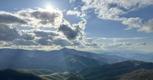 A panoramic view of rolling mountains under a bright sky with clouds and sunlight streaming through.