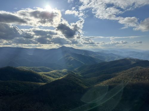 A panoramic view of rolling mountains under a partly cloudy sky, with sunlight illuminating the green valleys below.