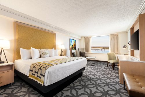 A modern hotel room featuring a king-sized bed, desk, armchair, and large window with a view.