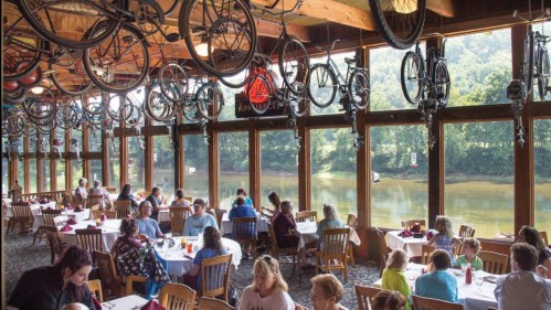 A lively restaurant with large windows overlooking a lake, decorated with bicycles hanging from the ceiling.