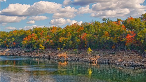 A serene lake bordered by vibrant autumn trees under a blue sky with fluffy clouds.