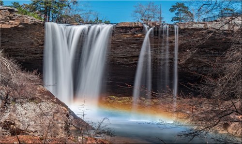 A serene waterfall cascades over rocky cliffs, surrounded by trees and a rainbow in the mist below.