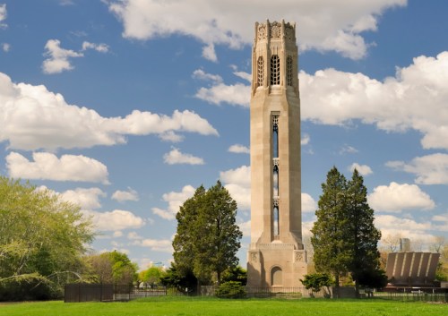 A tall stone tower surrounded by trees, set against a blue sky with fluffy white clouds.