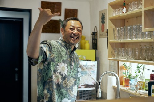 A smiling man in a floral shirt gives a thumbs-up gesture in a cozy kitchen filled with glassware and plants.