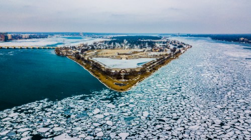 Aerial view of a snowy island surrounded by icy waters and floating ice, with a cloudy sky above.