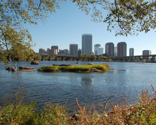 City skyline reflected in a river, with greenery and rocks in the foreground under a clear blue sky.