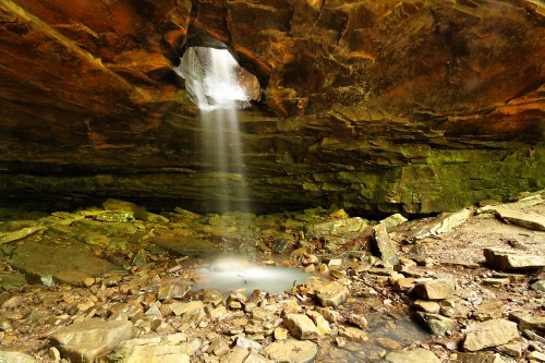 A serene cave with a waterfall cascading through a rock opening, surrounded by mossy stones and a tranquil pool below.