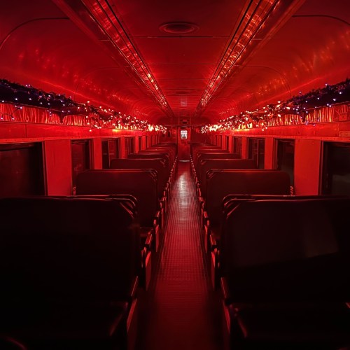A dimly lit train interior with red lighting and festive decorations along the ceiling. Empty rows of seats are visible.