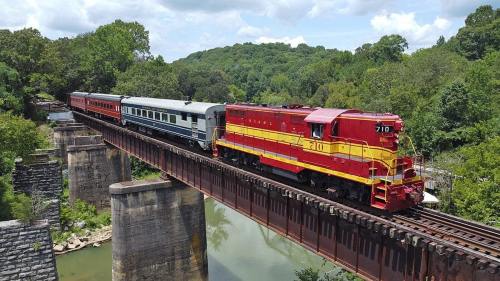 A red and yellow train crosses a bridge over a river, surrounded by lush green trees and a blue sky.