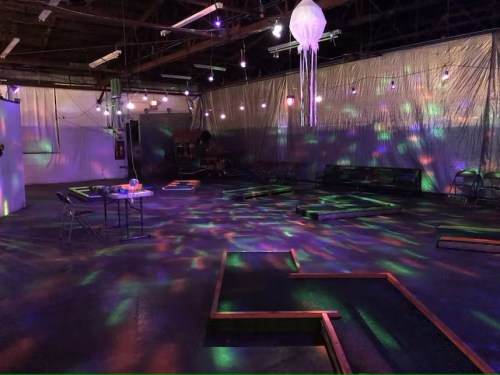 A dimly lit indoor space with colorful lights, featuring a mini-golf setup and a jellyfish decoration hanging from the ceiling.
