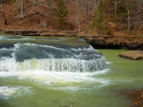 A serene waterfall cascades over a rocky ledge into a calm, green river surrounded by bare trees.