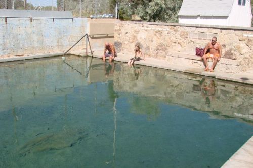 Three people sit by a clear, shallow pool, with one child dangling their feet in the water on a sunny day.