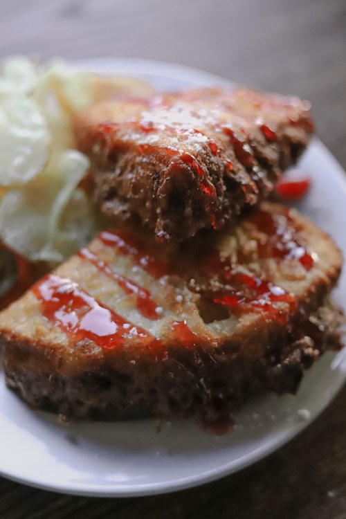 Close-up of two crispy, golden-brown sandwiches drizzled with ketchup, served with a side of pickles.