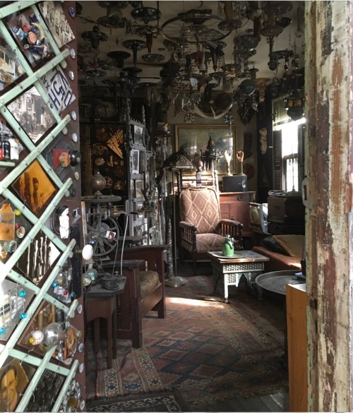 A cozy, eclectic room filled with vintage furniture, decor, and various hanging objects, illuminated by soft light.