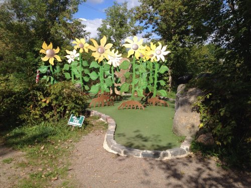 Colorful, oversized flower sculptures in a mini-golf setting, surrounded by greenery and a stone pathway.