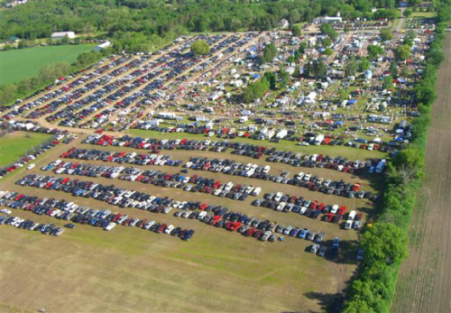 Aerial view of a large outdoor event with rows of parked cars and tents, surrounded by greenery and fields.