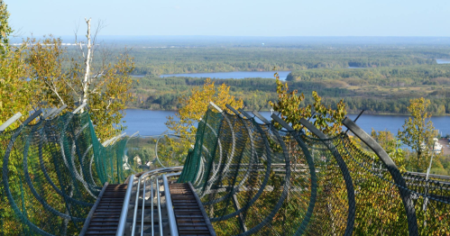 A scenic view from a hilltop, featuring a winding track and lush greenery overlooking a river and distant landscape.