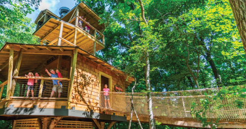 A wooden treehouse surrounded by trees, with people joyfully waving from different levels and a rope bridge connecting to it.