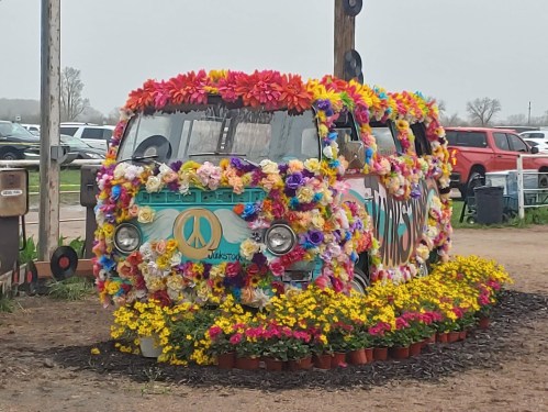 A colorful, flower-covered van with a peace sign, surrounded by potted flowers in a grassy area.