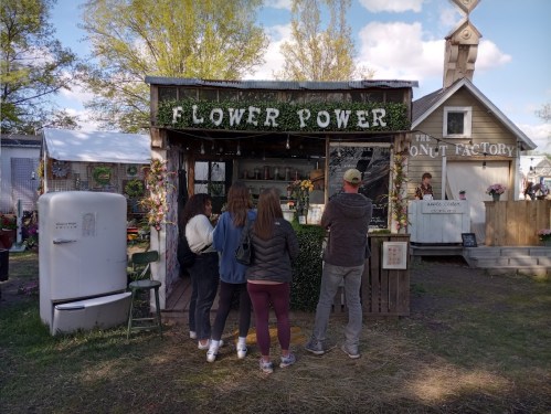 A group of four people stands in front of a flower shop called "Flower Power" in a vibrant outdoor setting.