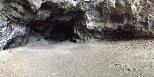A rocky cave entrance with a sandy floor, surrounded by rugged stone walls and shadows inside.
