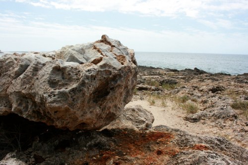 A large rock sits on a rocky beach, with the ocean and cloudy sky in the background.