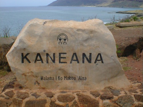 A large stone sign reading "Kaneana" with the phrase "Malama I Ko Makou Aina" near a coastal landscape.