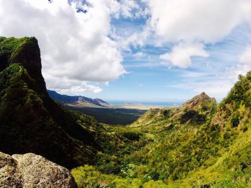 A panoramic view of a lush green valley surrounded by mountains, leading to a distant ocean under a partly cloudy sky.