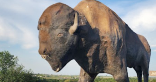 A large bison statue stands against a blue sky, surrounded by greenery.