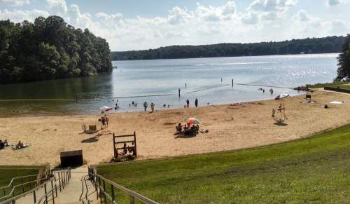 A sandy beach by a lake with people swimming, lounging under umbrellas, and trees in the background on a sunny day.