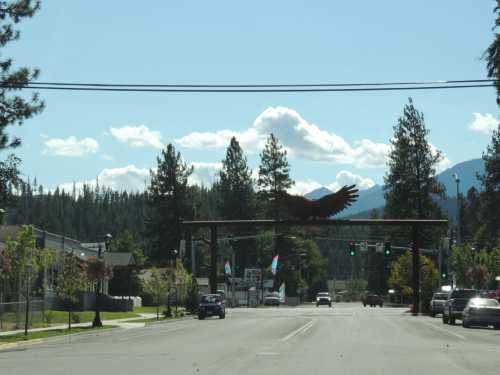 A street view featuring a large eagle archway, surrounded by trees and mountains under a clear blue sky.