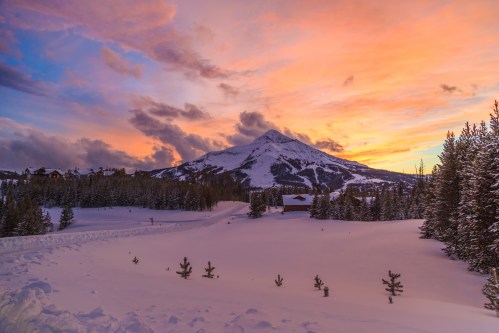 A snowy landscape at sunset, featuring a mountain silhouette and colorful clouds in the sky. Pine trees line the foreground.