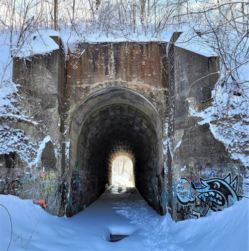 A snow-covered tunnel entrance with graffiti on the walls, leading to a bright light at the far end.