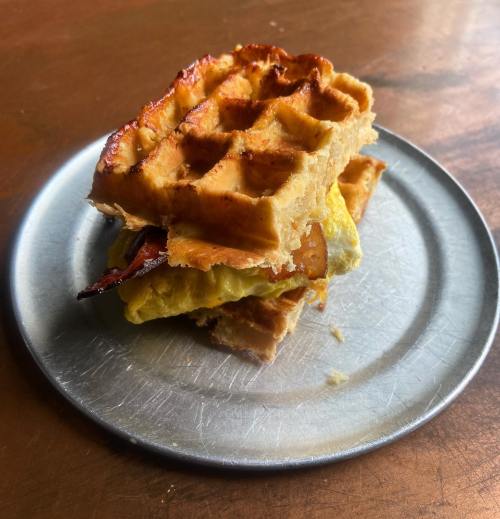 A stacked waffle sandwich with bacon and scrambled eggs on a metal plate.