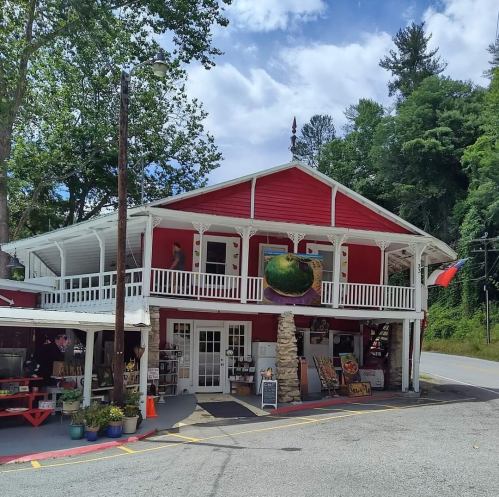 A red two-story building with a porch, featuring art displays and greenery, set against a blue sky and trees.