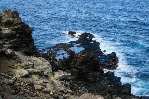 Rocky coastline with dark rocks and pools of water, surrounded by deep blue ocean waves.