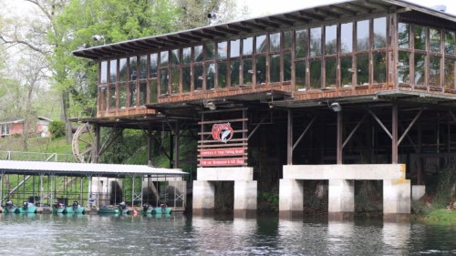 A rustic building with large windows sits over water, surrounded by greenery, with a sign displaying its name.
