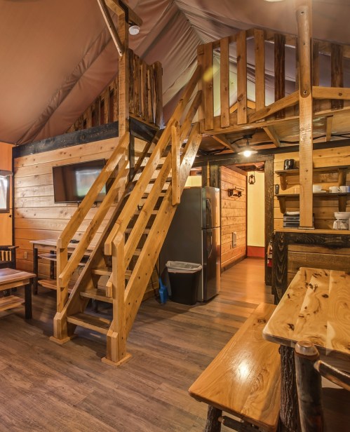 Interior of a cozy cabin featuring wooden stairs, a kitchen area, and rustic furniture. Warm lighting enhances the ambiance.