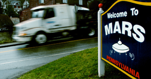A blurred truck passes by a colorful "Welcome to Mars, Pennsylvania" sign featuring a UFO design.