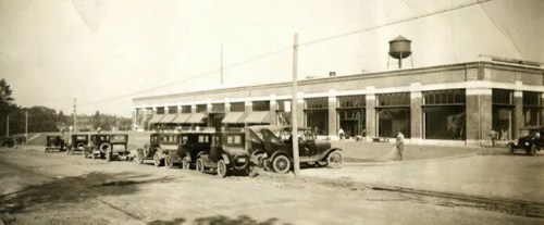 Historic black-and-white photo of early 20th-century cars parked outside a large building with a water tower.