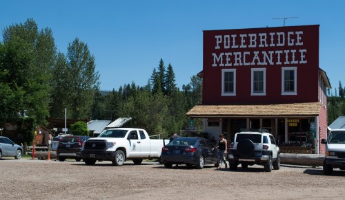 A rustic mercantile building with "Polebridge Mercantile" sign, surrounded by parked cars and trees under a clear blue sky.