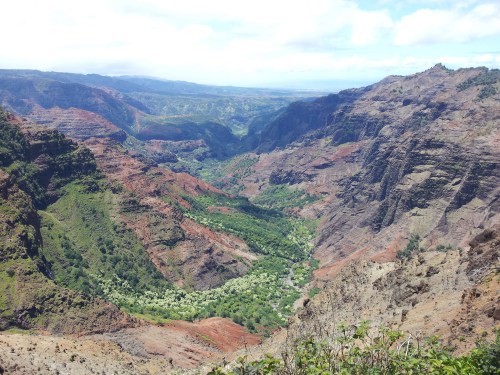 A panoramic view of a lush valley surrounded by steep, colorful cliffs under a partly cloudy sky.