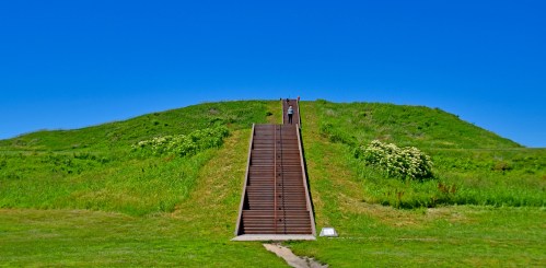A person walks up a set of stairs leading to a grassy hill under a clear blue sky.