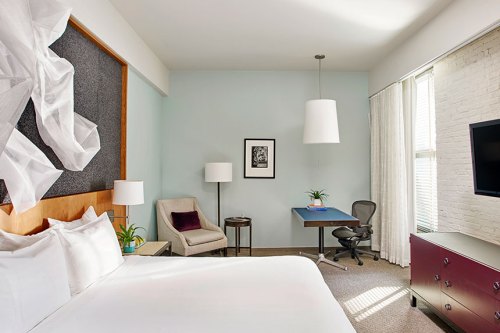 A modern hotel room featuring a bed, desk, chair, lamp, and large window with natural light.