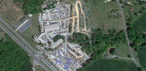 Aerial view of a large outdoor market with numerous vehicles and stalls, surrounded by greenery and roads.